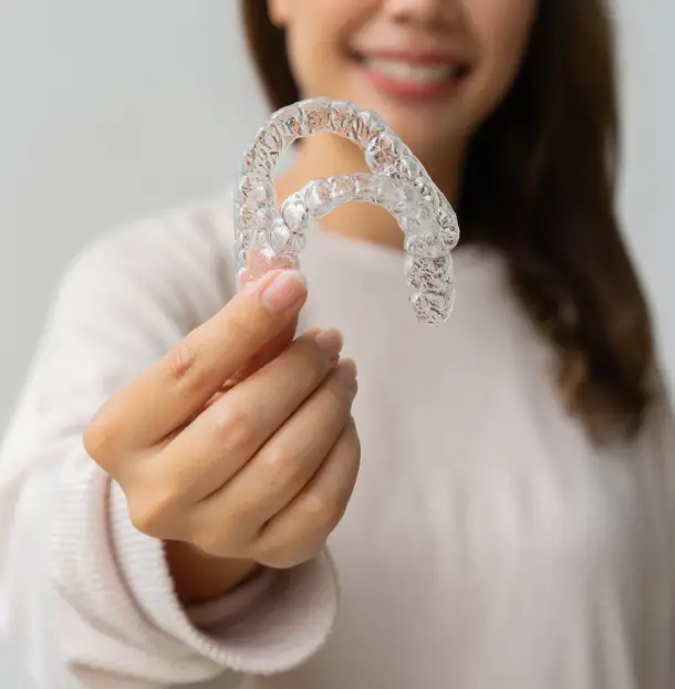 Woman holding clear dental aligner retainer toward camera in clinical white setting.