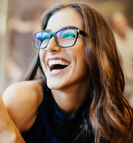 Smiling woman wearing glasses and black top in bright, blurred indoor setting.