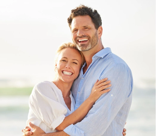 Happy couple hugging on beach in casual blue and white clothing, smiling at camera.
