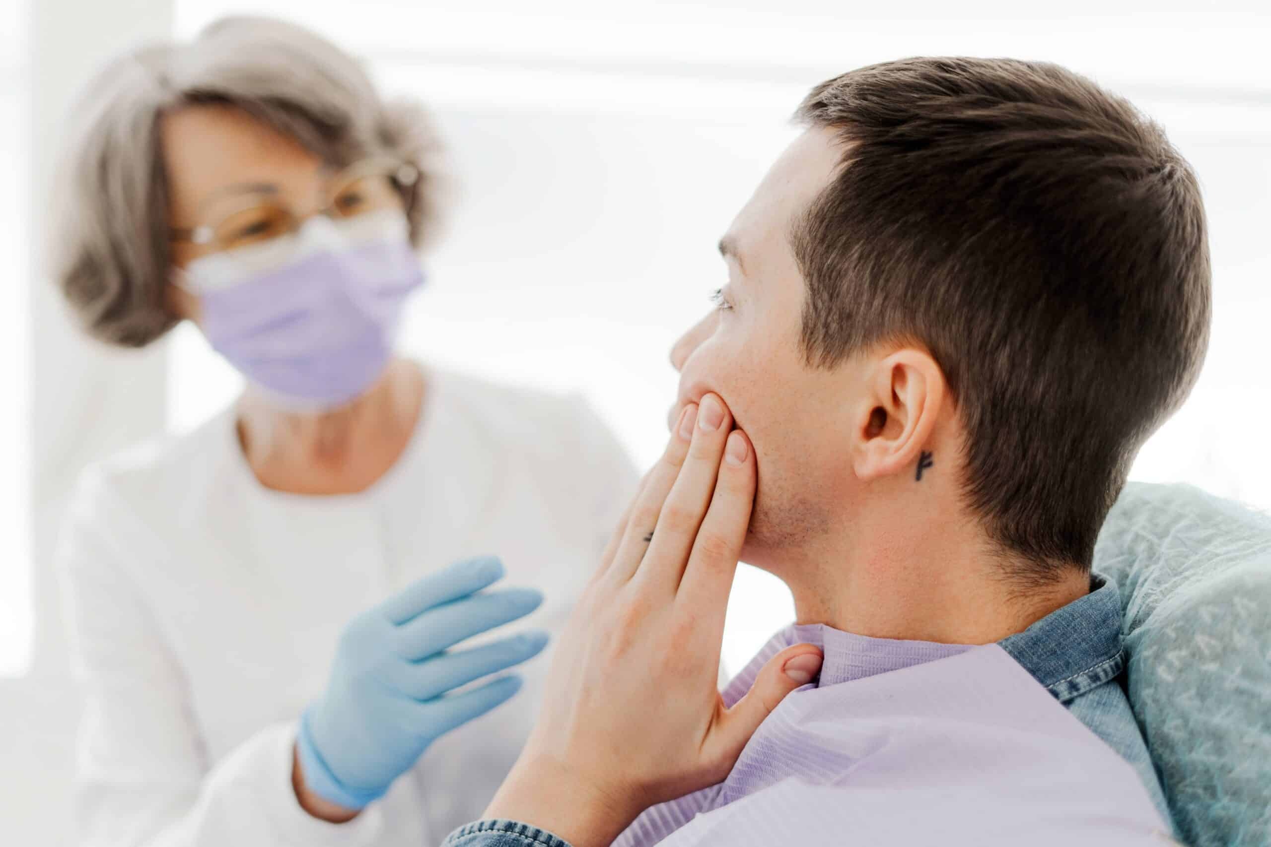 Male patient holding jaw in pain while dentist in mask examines him.